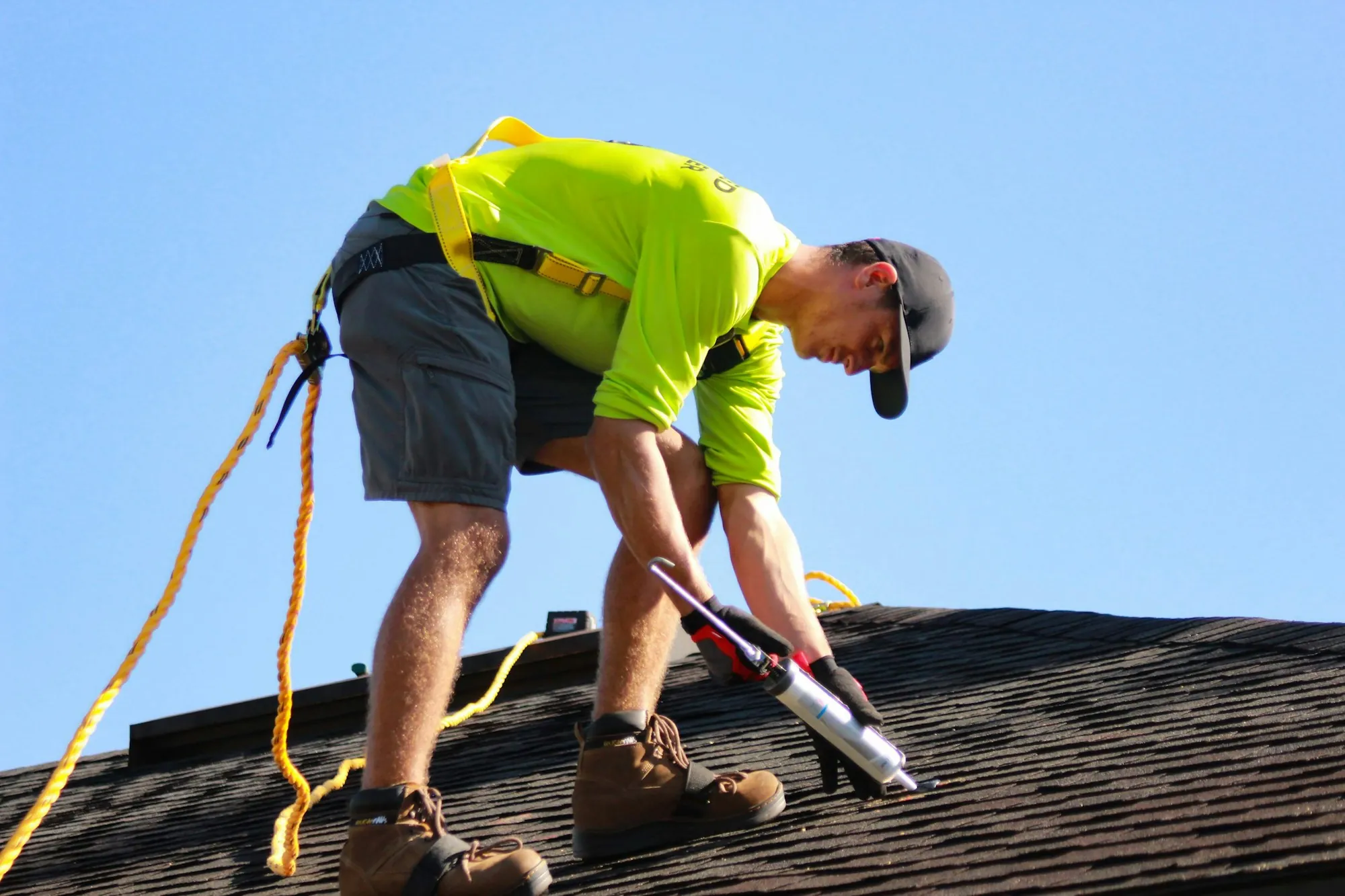 Roofer in a hi-vis shirt and harness applying sealant along a shingle roof